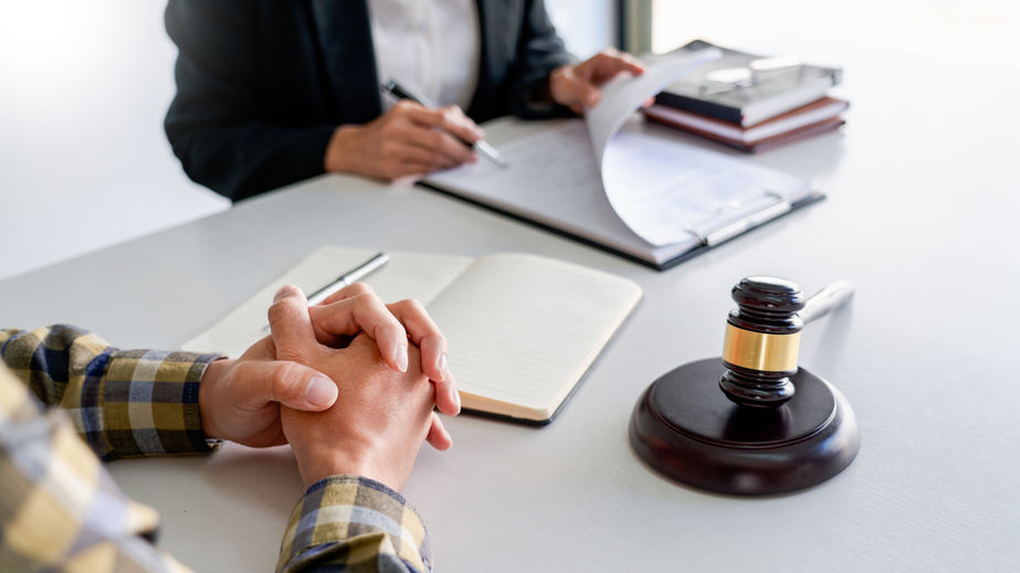 A legal consultation in progress with a lawyer flipping through documents and a client sitting opposite, hands clasped near a gavel and legal books on the table.