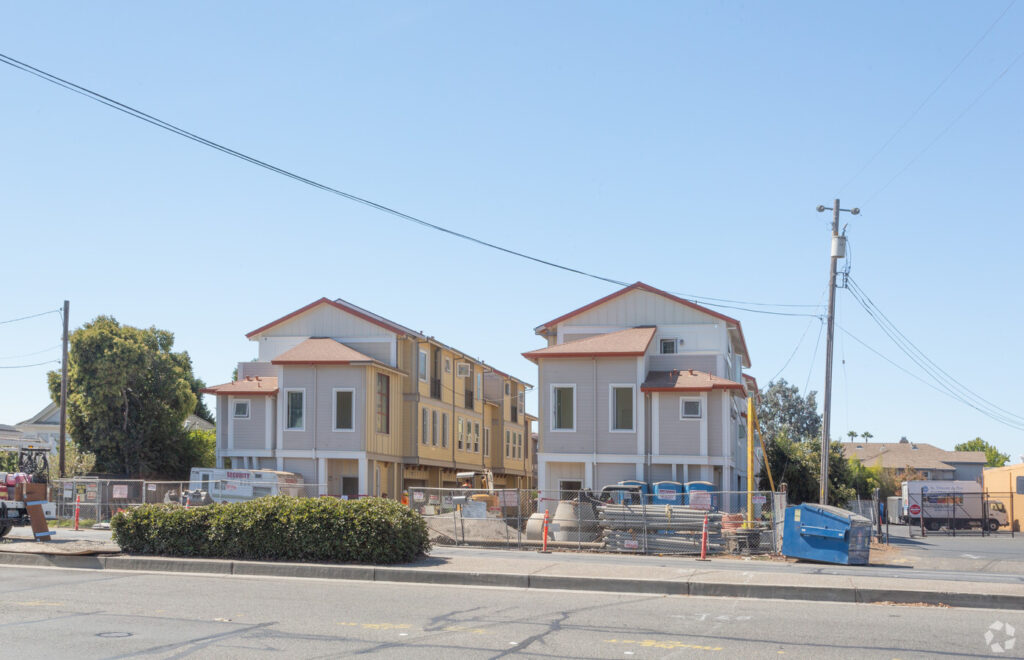 Construction site with multiple three-story residential buildings in progress. The area is fenced off, with construction equipment and materials visible. Clear blue sky in the background.