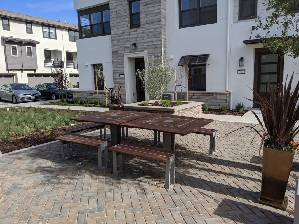 Outdoor seating area with wooden tables and benches on a brick patio in front of a modern residential building. Plants and a parked car are visible in the background.